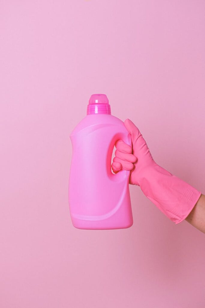 Unrecognizable person in pink protective glove demonstrating plastic bottle of detergent for daily cleaning against pink background in modern studio